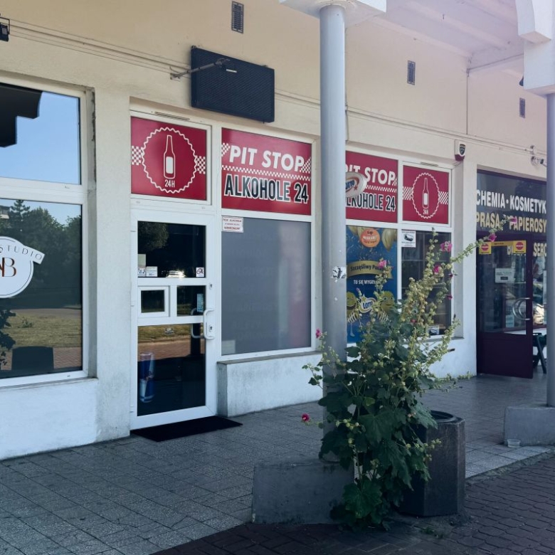 Row of shopfronts with red PIT STOP ALKOHOL 24 signs above glass doors and a potted plant in the foreground.