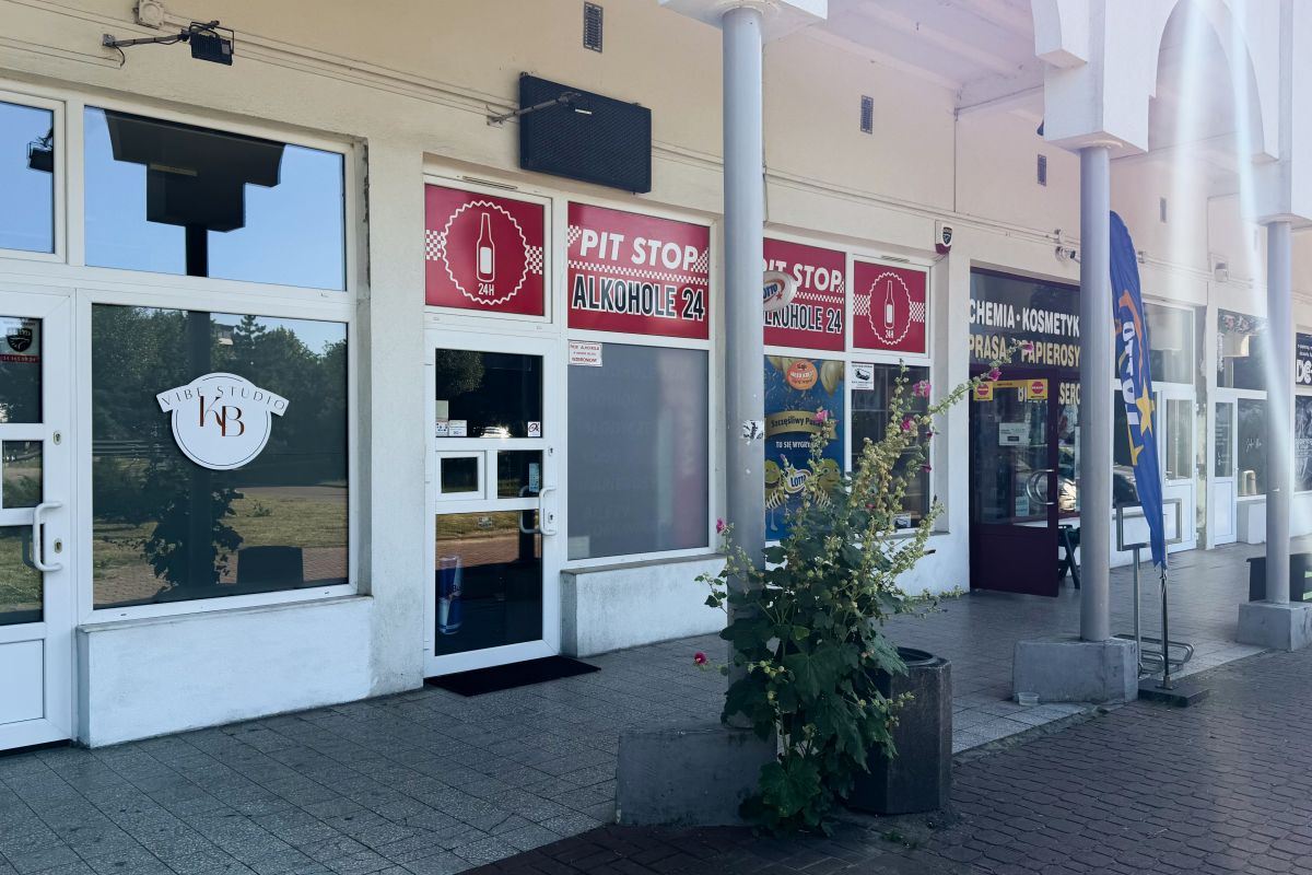 Row of shopfronts with red PIT STOP ALKOHOL 24 signs above glass doors and a potted plant in the foreground.