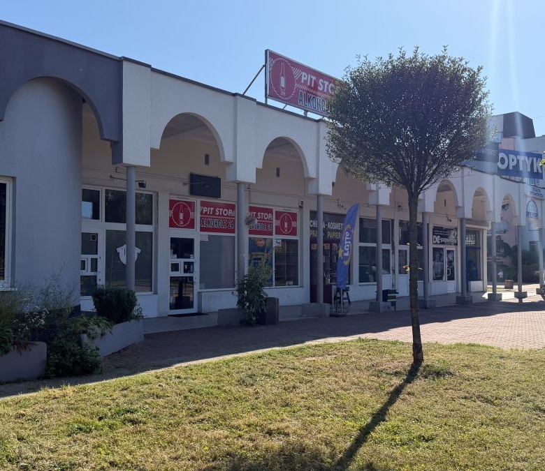 Row of storefronts with white arches along a pedestrian walkway under a sunny blue sky, some signs visible above the doors, and a parked car to the right.
