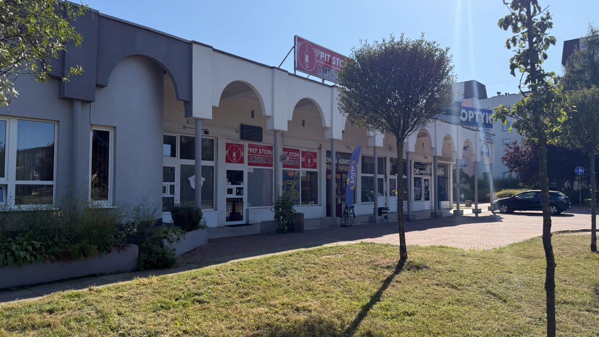Row of storefronts with white arches along a pedestrian walkway under a sunny blue sky, some signs visible above the doors, and a parked car to the right.
